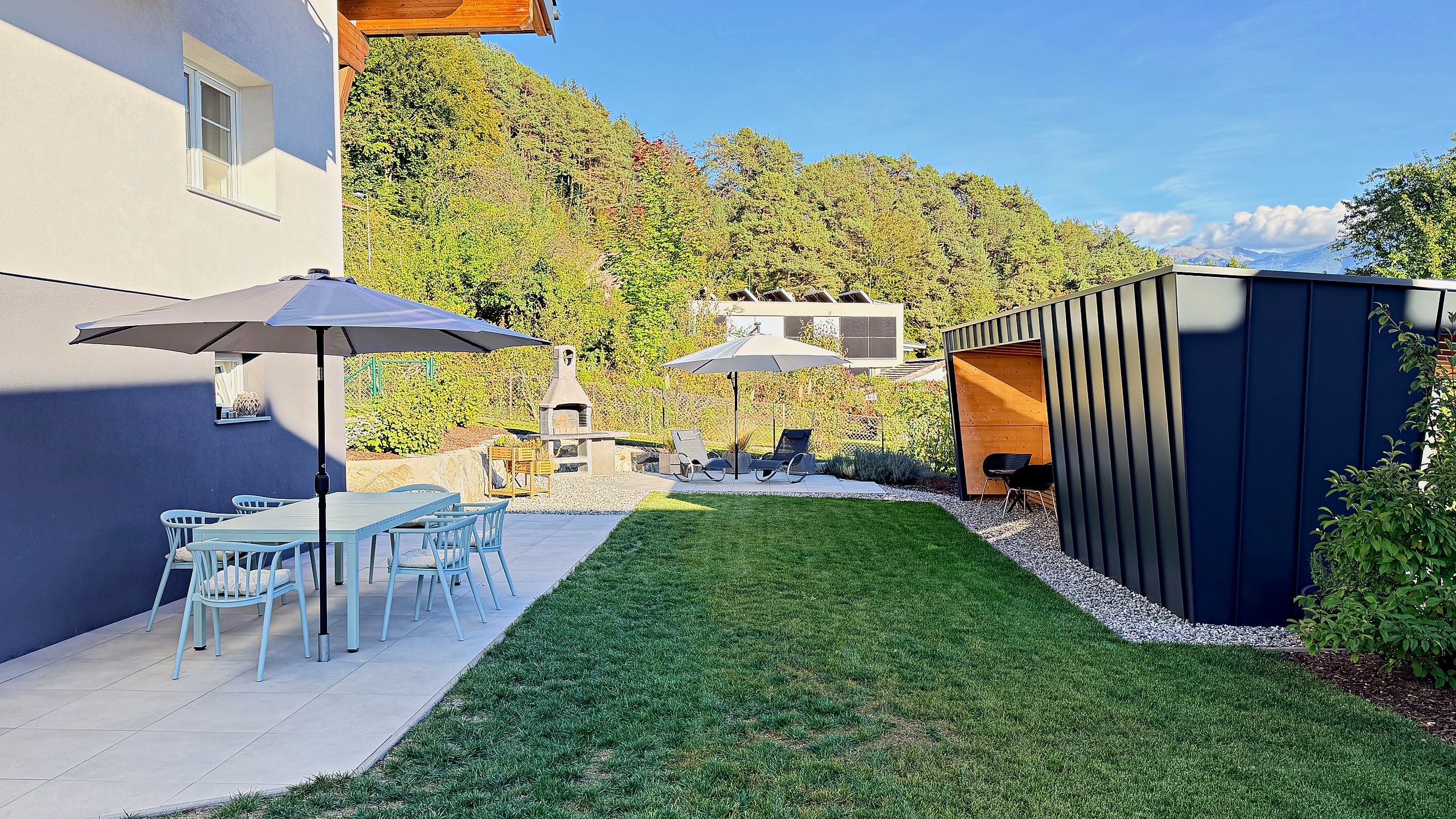 Terrasse mit Esstisch, Stühlen und Sonnenschirm direkt am Haus, daneben der Garten